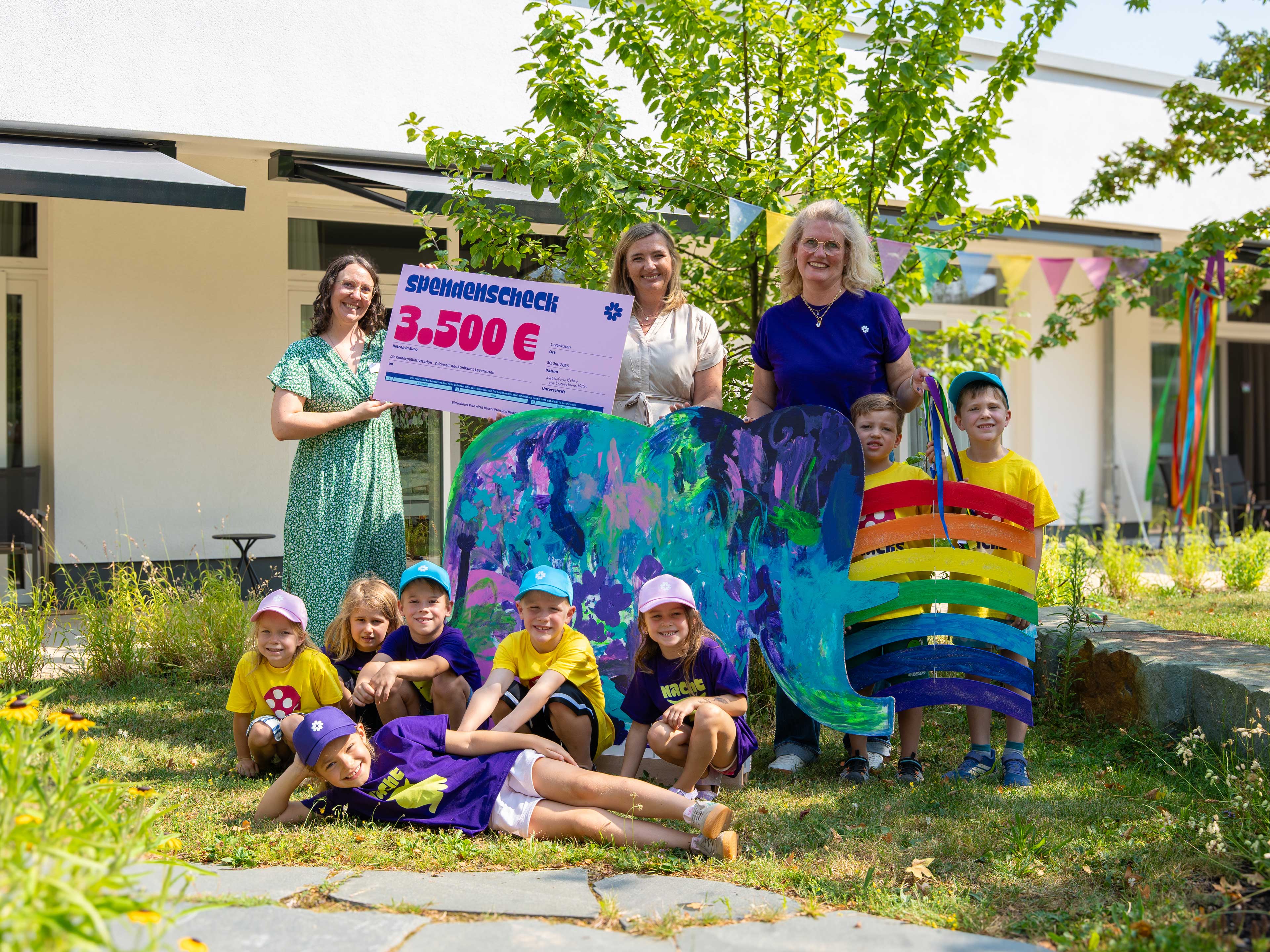 Agnes Busch, Geschäftsführerin von Katholino, überreicht gemeinsam mit Kindern der Kath. Kita St. Andreas den Spendenscheck und den beim Katholino-Sommerfest gestalteten Elefanten an Simone Vogel, Stationsleitung der Kinderpalliativstation „Zeitinsel“.